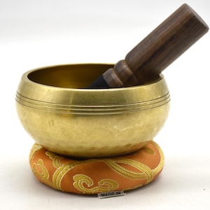 May include: A gold-colored metal singing bowl with a hammered texture and three horizontal etched lines around the rim. A dark brown wooden mallet rests inside the bowl. The bowl sits on a round, orange fabric cushion with gold patterns.