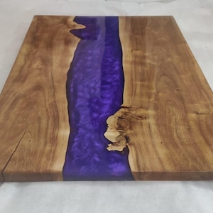 May include: A rectangular wooden serving board featuring a central river-like inlay of translucent, deep purple epoxy resin. The natural wood grain displays light brown tones with visible knots and organic edges along the resin section.