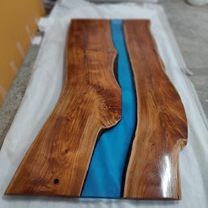 May include: A rectangular live edge wood table top featuring a central river design made of smooth, vibrant blue resin. The wood displays a rich, warm brown tone with visible natural grain patterns and knots, finished with a glossy, reflective surface.