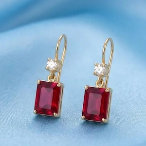 Puede incluir: Un par de pendientes colgantes de oro con piedras preciosas rojas rectangulares suspendidas de pequeñas piedras redondas transparentes. Los pendientes tienen un delicado mecanismo de gancho de hilo de oro y un diseño clásico y simétrico.