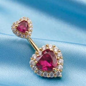 May include: A gold-toned navel piercing jewelry piece featuring two heart-shaped red stones. The smaller top heart and larger bottom heart are each surrounded by a halo of small, clear, round-cut white stones set in a metallic frame.