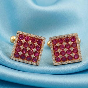 Puede incluir: Un par de gemelos cuadrados de oro con una cuadrícula de piedras redondas de color rojo vibrante. Cada gemelo está bordeado por un marco fino de piedras pequeñas, claras y brillantes, creando un diseño estructurado y simétrico.