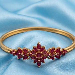 May include: A gold hinged bangle bracelet featuring a central floral-inspired cluster of marquise-cut and square-cut red gemstones. The decorative centrepiece extends along the band with symmetrical leaf-like arrangements of smaller red stones.