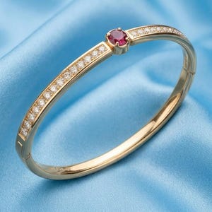 May include: A gold hinged bangle bracelet featuring a single oval-cut red ruby set in the center, flanked by a row of small round white diamonds embedded along the top half of the band, showcasing a polished metallic finish and a secure clasp.