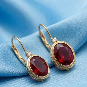 Puede incluir: Un par de pendientes de oro con cierre de palanca que presentan gemas ovaladas de color rojo intenso. Cada pendiente está acentuado con un pequeño diamante transparente de talla redonda sobre la piedra central, enmarcada por un borde dorado texturizado.