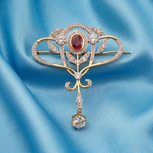 Pode incluir: Um broche de ouro com uma pedra preciosa vermelha de corte oval ao centro, rodeada por um halo de pequenos diamantes brancos. A estrutura apresenta um trabalho metálico intrincado, detalhes adicionais em diamante e um diamante pendente.