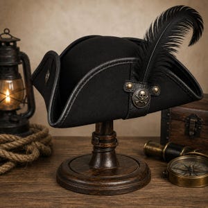 May include: A black tricorn hat featuring a prominent black feather plume and a circular metal emblem displaying a skull and crossbones design. The hat has a structured brim with dark trim and is displayed on a wooden stand.