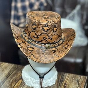 May include: A cowboy hat featuring a brown and black snakeskin pattern. The hat includes a black braided hatband adorned with a circular metallic emblem at the centre and a brown chin strap with an adjustable toggle for a secure fit.