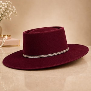 Puede incluir: Un sombrero de fieltro color burdeos intenso con copa plana y ala ancha y rígida. El sombrero presenta un cordón fino de color dorado metálico envuelto alrededor de la base de la copa, atado con un pequeño detalle decorativo en el centro frontal.