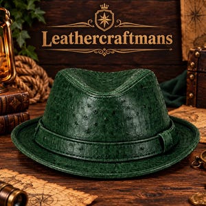 May include: A dark green fedora hat with a textured ostrich skin pattern. The hat features a matching green band around the base of the crown and a pinched top. The text Leathercraftmans is visible in the background above the hat.