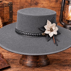 Könnte beinhalten: Ein breitkrempiger grauer Hut mit flacher Krone, der mit einem schwarzen geflochtenen Lederband um die Basis versehen ist. Eine dekorative beige Blume mit dunklen Blättern und kleinen Metallnieten ist seitlich am Band befestigt.