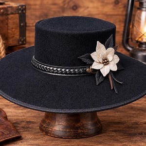 May include: A black flat-brimmed hat featuring a braided black leather band around the crown. A decorative accent on the side consists of a cream-coloured flower with dark leaves and small metallic beads at the centre of the floral arrangement.
