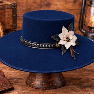 Puede incluir: Un sombrero azul marino de ala plana con una banda de cuero trenzado negro alrededor de la copa. El sombrero está decorado con una gran flor de color crema de varias capas, acompañada de hojas de color verde oscuro y pequeños tachones metálicos.