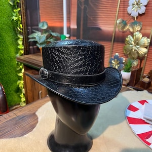 May include: A black top hat with a textured crocodile skin pattern. The hat features a black band around the base of the crown, accented with a silver-toned metal buckle on the side. The hat is displayed on a smooth, featureless black mannequin head.