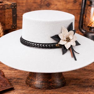 May include: A light grey flat-brimmed hat featuring a black braided leather band around the crown. The hat is decorated with a layered floral accent made of cream and black petals, centred with small metallic beads and dark green leaves.