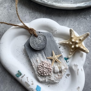 May include: A grey, pentagonal hanging ornament with a textured surface featuring a small beige starfish, a round pink coral piece, two white pebbles, and a circular grey medallion with a lion emblem and the text "SUOMEN LEIJONA".