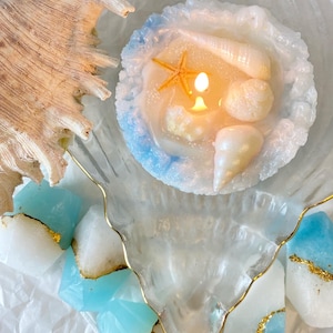 Puede incluir: Una vela redonda blanca con borde azulado, que presenta formas incrustadas de estrellas de mar y conchas marinas, se encuentra encendida dentro de un plato transparente con forma de concha y borde dorado, rodeada de piezas de cera geométricas.