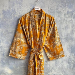 Può includere: Una vestaglia color giallo senape con una stampa floreale e botanica bianca su tutta la superficie. Il capo presenta maniche lunghe e ampie, una cintura in vita coordinata e profili bianchi lungo l'apertura frontale e i polsini. È presente un'etichetta con la scritta IMPEROX.