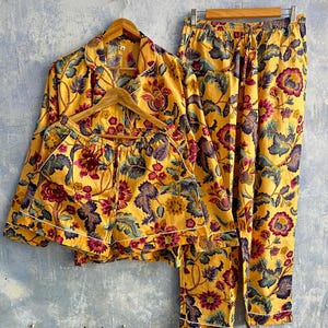 May include: A yellow pajama set featuring a long-sleeve button-down shirt, matching shorts, and long pants, all decorated with a vibrant floral pattern in shades of magenta, purple, and teal, finished with white piping along the edges.