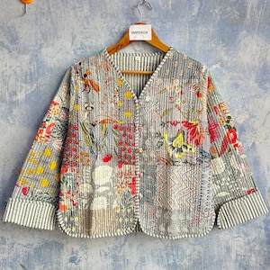Puede incluir: Una chaqueta acolchada de manga larga con un diseño de patchwork que presenta varios patrones florales y geométricos en tonos gris, rojo, amarillo y blanco. La chaqueta tiene cuello en V, cierre frontal de botones y puños con rayas blancas y negras.