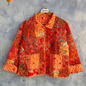 May include: A quilted jacket featuring a vibrant orange and red floral patchwork pattern. The garment includes a collar, long sleeves with striped cuffs, and two front pockets with white and orange vertical stripes and small red heart details.
