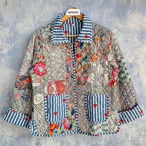 Op de afbeelding: Een gewatteerd jack met een patchwork van grijze bloemen- en paisley-patronen, geaccentueerd met blauw-witte gestreepte biezen op de kraag, manchetten en twee voorzakken. Kleine rode hartmotieven zijn verspreid over de stof en de zakken.