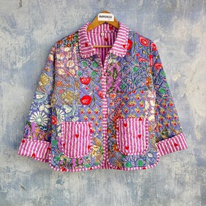Peut inclure: Une veste matelassée au design patchwork avec des motifs floraux dans des tons de violet, bleu et rouge. Le vêtement comprend un col, des poignets et deux poches avant à rayures verticales blanches et violettes avec de petits cœurs rouges.