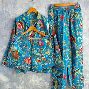 Op de afbeelding: Een blauwe pyjamaset met een levendig bloemen- en paisleypatroon in rood-, geel- en wittinten. De set bestaat uit een overhemd met lange mouwen en knopen, een bijpassende korte broek en een lange broek met een elastische tailleband.