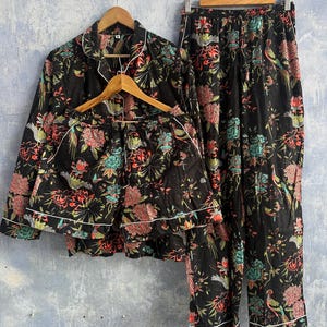 Puede incluir: Un conjunto de pijama negro que incluye una camisa de manga larga con botones, pantalones cortos y pantalones largos a juego. La tela presenta un estampado floral y de aves en tonos rojo, verde y blanco, con ribetes blancos en los bordes.