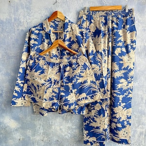 Op de afbeelding: Een blauwe pyjamaset bestaande uit een pyjamashirt met lange mouwen en knopen, bijpassende shorts en een lange broek, allemaal voorzien van een beige bloemenpatroon. Elk kledingstuk heeft witte biezen langs de randen en elastische taillebanden.