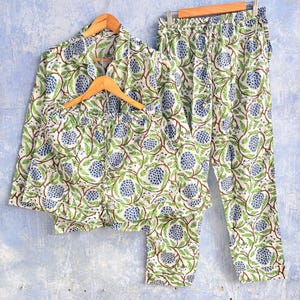 Op de afbeelding: Een pyjamaset bestaande uit een shirt met lange mouwen en knoopsluiting, een lange broek en een korte broek. De kledingstukken hebben een witte basis met een herhalend patroon van blauwe bloemtrossen en groene ranken met bruine stengels.