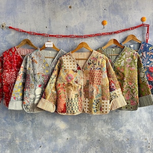 May include: Five quilted jackets with V-necks and long sleeves, each featuring distinct floral and geometric block-print patterns in vibrant colors including red, yellow, green, and blue, all displayed on wooden hangers against a textured wall.