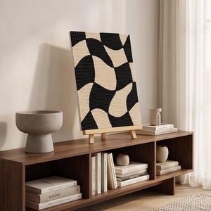 May include: A rectangular canvas featuring a black and beige wavy checkerboard pattern displayed on a wooden easel. The artwork is composed of a dense grid of small dots, creating a textured appearance with alternating curved shapes in black and beige.