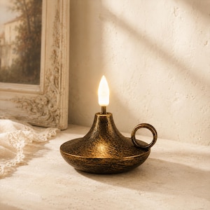 May include: A low-profile oil lamp featuring a dark, distressed bronze-toned finish with a circular handle on the side. The lamp is topped with a single, upright, glowing white flame-shaped bulb that emits a soft, warm light from its center.