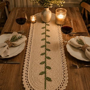 May include: An oval-shaped cream-colored crochet table runner featuring a central green vine pattern with leaves. The runner has a decorative scalloped border and a green tassel at the bottom end, providing a textured accent for a dining table.