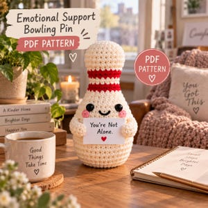 Op de afbeelding: Een crèmekleurige gehaakte bowlingkegel met een rode streep rond de nek, zwarte ogen en roze wangen. Het figuurtje houdt een wit bordje vast met de tekst "You're Not Alone." en een klein rood hartje. De tekst "PDF PATTERN" is duidelijk zichtbaar.