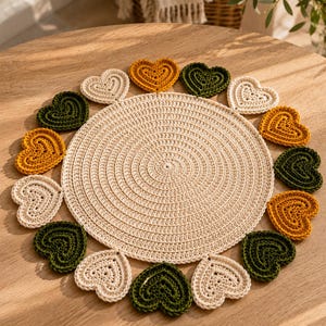 May include: A circular crochet placemat in cream yarn featuring a central flat disc surrounded by a border of alternating heart-shaped motifs in cream, mustard yellow, and dark forest green colors, all crafted with a consistent textured stitch pattern.