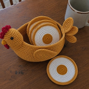 May include: A mustard yellow crocheted chicken-shaped basket containing several round coasters with white and mustard yellow centers. One matching round coaster lies flat outside the basket. The chicken has a red comb and wattle.