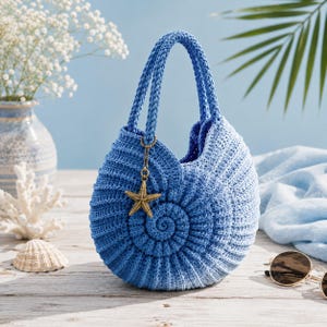 May include: A round, blue crocheted handbag shaped like a nautilus shell, featuring a gradient colour effect from dark to light blue. The bag has two braided handles and a gold-toned metal starfish charm attached to one of the handle rings.