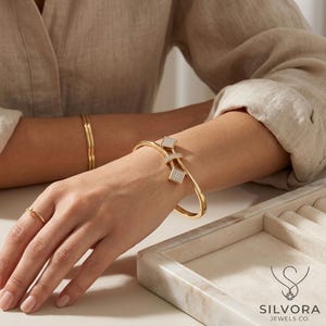May include: A gold-toned hinged bangle bracelet featuring two square-shaped charms encrusted with small clear stones. The bracelet is worn on the wrist alongside a thin gold-toned band and a simple gold ring. The logo for Silvora Jewels Co. is visible.