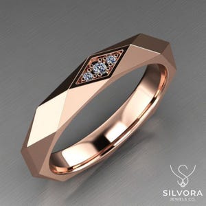 May include: A rose gold ring featuring a faceted geometric band with a central diamond-shaped inlay. The inlay contains three small, clear, round-cut stones arranged in a vertical line. The ring has a polished finish and a sharp, angular design.