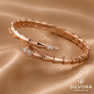 Puede incluir: Una pulsera con forma de serpiente en tono oro rosa con un diseño segmentado y flexible. Las secciones de la cabeza y la cola están adornadas con pequeñas piedras claras y brillantes. El nombre SILVORA JEWELS CO. aparece impreso en blanco.