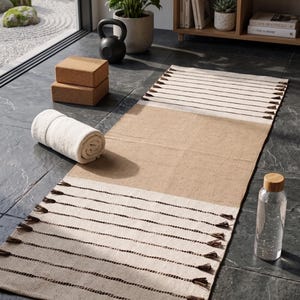 Op de afbeelding: Een rechthoekige yogamat met een centraal beige gedeelte en twee gebroken witte uiteinden met donkerbruine horizontale strepen en decoratieve kwastjes. De mat wordt vergezeld door twee kurkblokken, een opgerolde witte handdoek en een waterfles.