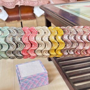 May include: A collection of eleven wavy, textured bangles arranged in a row on a clear rod. The bangles feature a repeating rippled pattern and come in a variety of colours including light green, grey, red, cream, yellow, brown, and dark red.