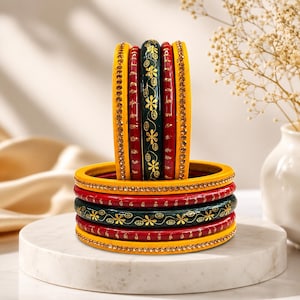May include: A set of five circular bangles featuring a vibrant colour palette of yellow, red, and dark green. The yellow bangles are adorned with small embedded crystals, while the dark green bangle displays a delicate gold floral pattern.
