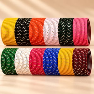 Op de afbeelding: Twee stapels dunne metalen armbanden gerangschikt in een horizontale rij. De bovenste stapel bevat gele, zwarte, oranje, witte, donkergroene en roze armbanden. De onderste stapel bevat groene, witte, roze, blauwe, gele en kastanjebruine armbanden met gouden stippen.