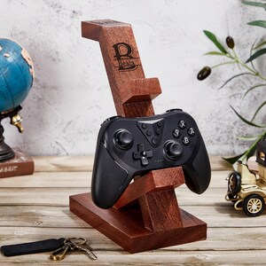 May include: A dark brown wooden gaming controller stand with a rectangular base and an angled support arm. The stand features a laser-engraved logo with the letter B and the name Beata. A black wireless gaming controller rests on the stand.