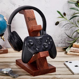 May include: A dark brown wooden stand featuring the name Vince engraved on the vertical support. The stand holds a pair of black over-ear headphones on the top arm and a black wireless video game controller on the lower front-facing arm.