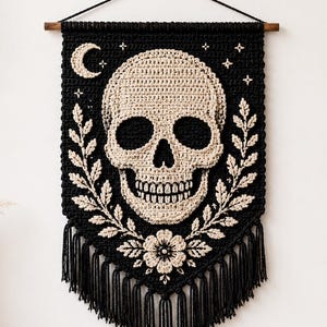 May include: A black and cream crochet wall hanging featuring a central skull motif surrounded by leafy branches and a flower at the bottom. The design includes a crescent moon and stars at the top, suspended from a wooden rod with black string.