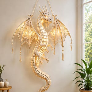 Puede incluir: Un tapiz de macramé color crema con forma de dragón, con alas intrincadamente anudadas y una cola larga y curva. El dragón cuenta con una fuente de luz cálida interna que ilumina su cuerpo y alas, suspendido de un anillo de latón circular.